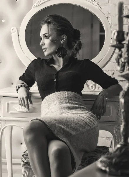 Молодая довольно элегантная серьезная женщина, в черном топе и легкой юбке, сидит в роскошной комнате с белым зеркалом позади нее и подсвечником на столе, в черно-белом — стоковое фото