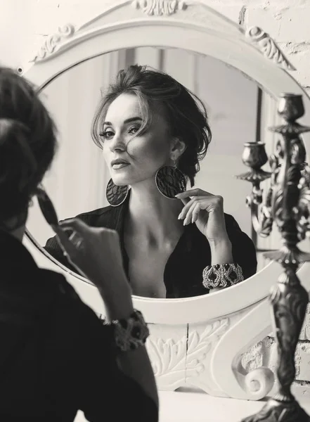 Närbild porträtt av ung ganska elegant allvarlig kvinna med vackra makeup, bär svart topp och ljus kjol, sitter i lyxrum med vita spegeln framför henne och ljusstake på bordet, i svart och vitt — Stockfoto
