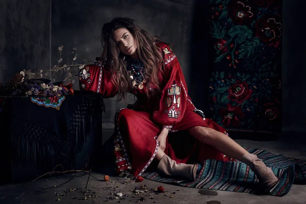 Красивая молодая сексуальная женщина с длинными каштановыми волосами, в красном платье с традиционным украинским орнаментом и светло-коричневыми сапогами, сидит на полу в темной студии — стоковое фото