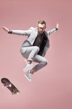 Gözlük, Kahverengi saç ve sakal, genç yakışıklı komik adam ışık gri takım elbise ve spor ayakkabı, kaykay ile hafif pembe arka plan atlama giyiyor 