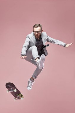 Işık gri takım elbise ve spor ayakkabı, kaykay ile hafif pembe arka plan atlama giyen genç yakışıklı ciddi adam gözlük, Kahverengi saç ve sakal, 