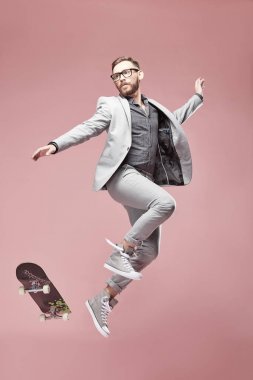 Gözlük, Kahverengi saç ve sakal, genç yakışıklı komik adam ışık gri takım elbise ve spor ayakkabı, kaykay ile hafif pembe arka plan atlama giyiyor 