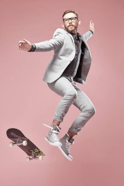 Genç yakışıklı ciddi adam gözlük, Kahverengi saç ve sakal ışık gri takım elbise ve spor ayakkabı giyen, kaykay ile atlama ve ışık üzerinde uçan pembe arka plan 