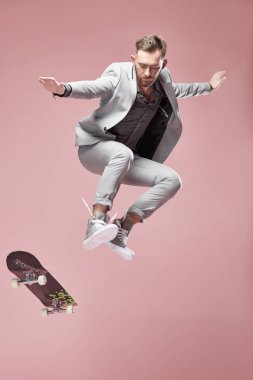 Genç yakışıklı ciddi adam gözlük, Kahverengi saç ve sakal ışık gri takım elbise ve spor ayakkabı giyen, kaykay ile atlama ve ışık havada uçan pembe arka plan 