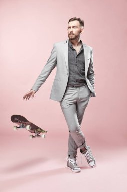 Işık gri takım elbise ve spor ayakkabı, açık pembe renkli üzerinde kaykay ile ayakta giyen genç yakışıklı ciddi adam gözlük, Kahverengi saç ve sakal, 