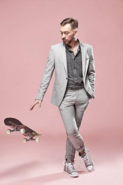 Kahverengi saç ve sakal, genç yakışıklı ciddi adam ışık gri takım elbise ve spor ayakkabı, açık pembe renkli üzerinde kaykay ile ayakta 