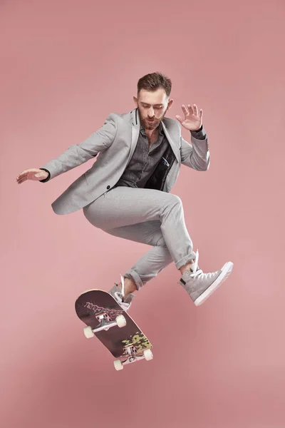 光の灰色のスーツとスニーカー、光のピンクの背景に、スケート ボードでジャンプを着て茶色の髪とひげ、若いハンサムな男 — ストック写真