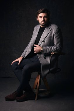 Güzel saç ve sakal ve ciddi bir yüz, yakışıklı seksi genç adam tahta sandalyede koyu gri Studio elinde gri ceket ve izlemek ile takım elbise poz