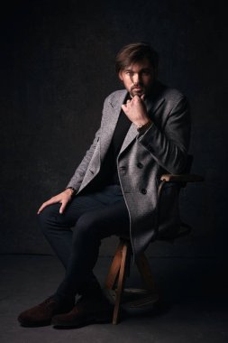 Güzel saç ve sakal ve ciddi bir yüz, yakışıklı seksi genç adam tahta sandalyede koyu gri Studio elinde gri ceket ve izlemek ile takım elbise poz