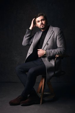 Yakışıklı genç adam tahta sandalyede koyu gri Studio elinde gri ceket ve izlemek ile takım elbise poz güzel saç ve sakal ve ciddi bir yüz,