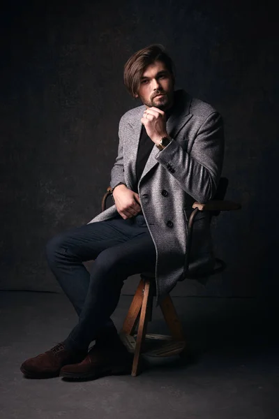 Удивительный сексуальный молодой человек с красивыми волосами и бородой и серьезным лицом, позирующий на деревянном стуле, в костюме с серой курткой и часами на руке, в темно-серой студии — стоковое фото