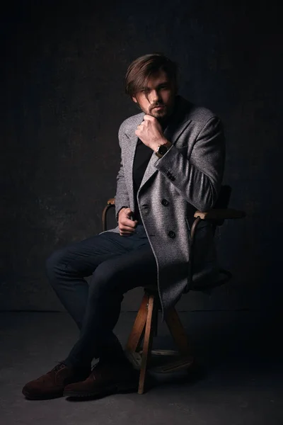 ハンサムなセクシーな若い男美しい髪とひげと深刻な顔、暗い灰色のスタジオで、彼の手でグレーのジャケットと時計とスーツを着て木の椅子のポーズ — ストック写真