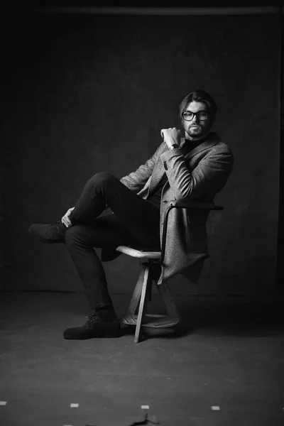 Amazing Sexig ung man med vackra hår och skägg och allvarliga ansikte, poserar på den trä stolen, klädd i kostym med grå jacka och titta på handen, i mörk grå studio, i svart och vitt — Stockfoto
