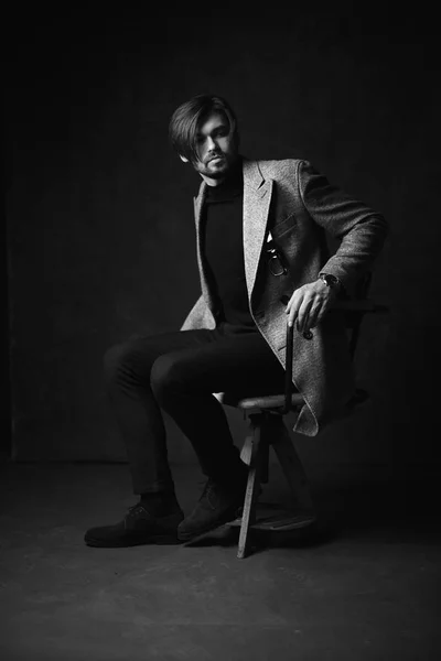 Amazing Sexig ung man med vackra hår och skägg och allvarliga ansikte, poserar på den trä stolen, klädd i kostym med grå jacka och titta på handen, i mörk grå studio, i svart och vitt — Stockfoto