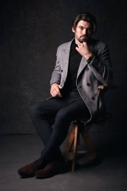 Güzel saç ve sakal ve ciddi bir yüz koyu gri Studio elinde gri ceket ve izlemek ile takım elbise ahşap sandalye üzerinde oturan, yakışıklı genç erkek modeli