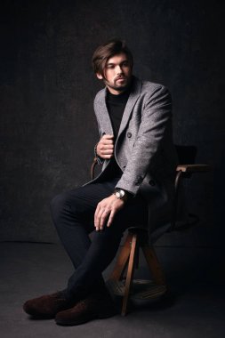 Güzel saç ve sakal ve ciddi bir yüz koyu gri studio, gri ceket, takım elbise giyen ahşap sandalye üzerinde oturan, yakışıklı genç erkek modeli