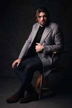 Yakışıklı genç adam tahta sandalyede koyu gri Studio elinde gri ceket ve izlemek ile takım elbise poz güzel saç ve sakal ve ciddi bir yüz,