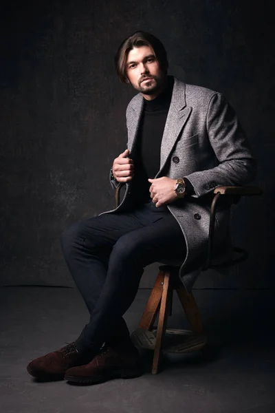 Handsome young male model with beautiful hair and beard, sitting on the wooden chair, wearing suit with grey jacket, in dark grey studio
