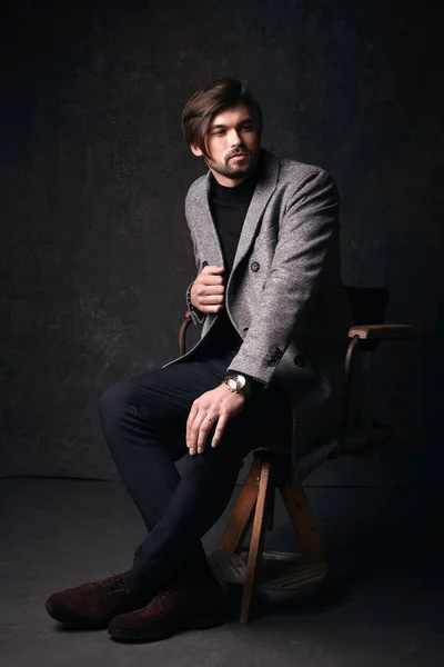 Handsome young male model with beautiful hair and beard and serious face, sitting on the wooden chair, wearing suit with grey jacket, in dark grey studio