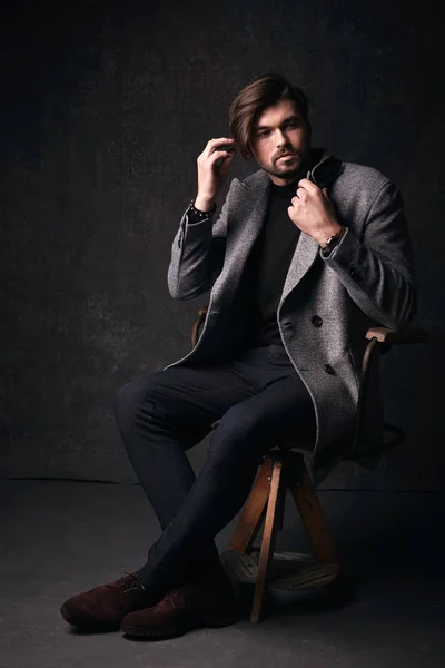Handsome young male model with beautiful hair and beard and serious face, posing on the wooden chair, wearing suit with grey jacket and watch on his hand, in dark grey studio