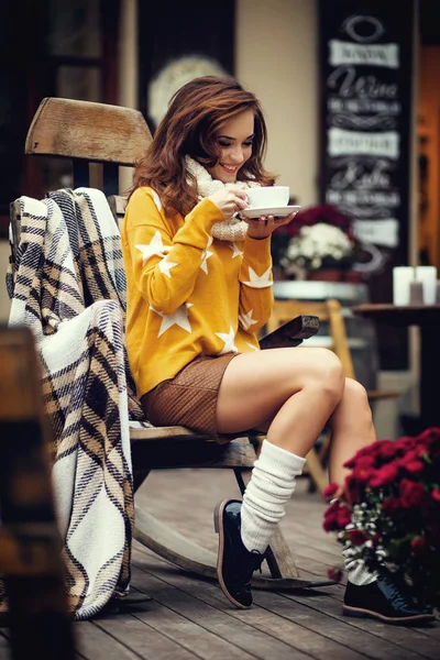 Hermosa joven morena con el pelo largo, sentada en la cafetería al aire libre, vistiendo suéter amarillo y bebiendo de la taza blanca — Foto de Stock