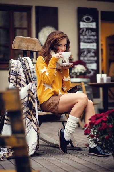 Hermosa joven morena con el pelo largo, sentada en la cafetería al aire libre, vistiendo suéter amarillo y bebiendo de la taza blanca — Foto de Stock