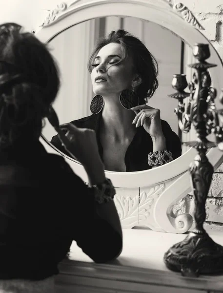 Портрет молодой привлекательной брюнетки с гламурным макияжем и в черной блузке с глубокой шеей, смотрящей и позирующей возле круглого зеркала в черно-белом — стоковое фото