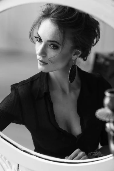 Portret młoda atrakcyjna kobieta brunetka czarujący makijaż i na sobie czarną bluzkę deep necked, patrząc i pozowanie w pobliżu okrągłe lustro biały czarno-biały — Zdjęcie stockowe