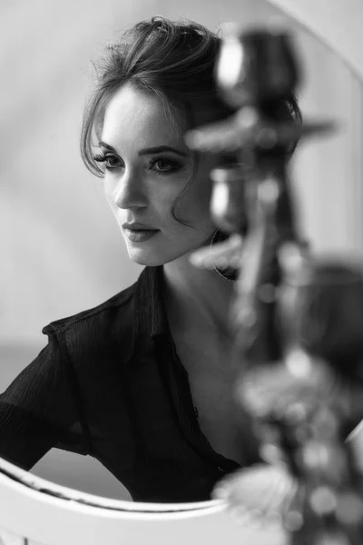 Porträtt av ung attraktiv brunett anbud kvinna med glamorös makeup och bär svart djup hals blus, tittar och poserar nära runda vita spegeln, i svart och vitt — Stockfoto