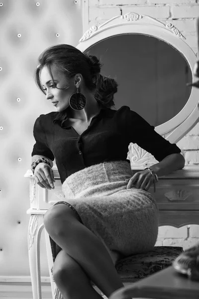 Porträtt av ung attraktiv brunett anbud kvinna med glamorös makeup, bär svart djup hals blus och ljus kjol, tittar ner och posing nära runda vita spegeln, i svart och vitt — Stockfoto
