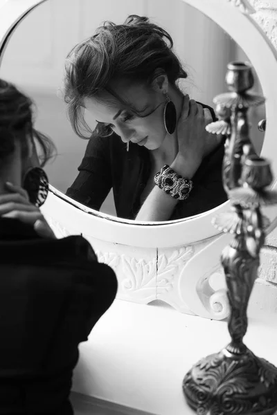 Portrait of young attractive brunette tender woman with glamorous makeup, wearing black deep-necked blouse and light skirt, looking down and posing near the round white mirror, in black and white — Stock Photo, Image