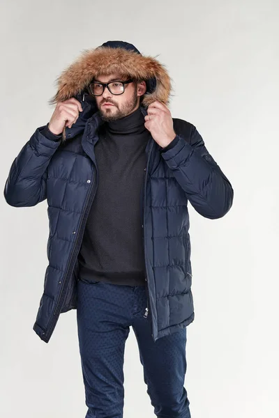 Attractive young sexy man model with beard and glasses posing in dark blue warm jacket with hood with a feather, and dark blue pants in light studio — Stock Photo, Image