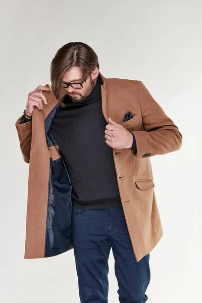 Attraktiv ung sexig man modellen med skägg och glasögon poserar i ljus brunt cashmere kappa och mörk blå byxor i ljus studio — Stockfoto