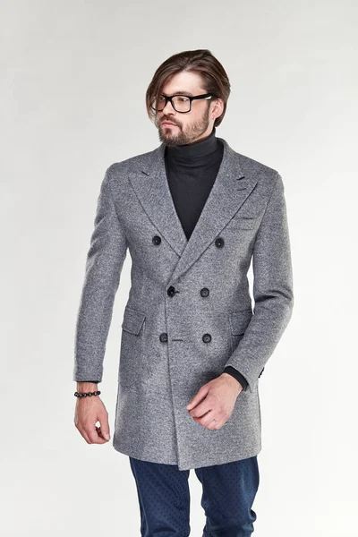 Atractivo joven sexy hombre serio modelo con barba y gafas posando en abrigo de cachemira gris y pantalones azul oscuro en estudio de luz —  Fotos de Stock