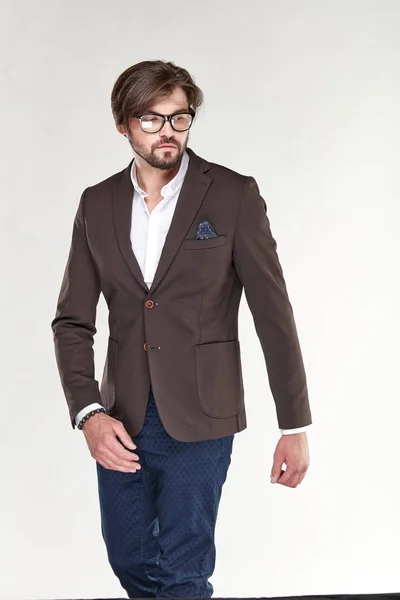 Attractive young sexy serious man model with beard and glasses posing in brown jacket, white shirt dark blue pants in light studio — Stock Photo, Image