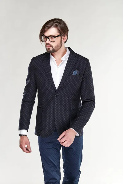 Attractive young sexy serious man model with beard and glasses posing in black jacket, white shirt dark blue pants in light studio — Stock Photo, Image