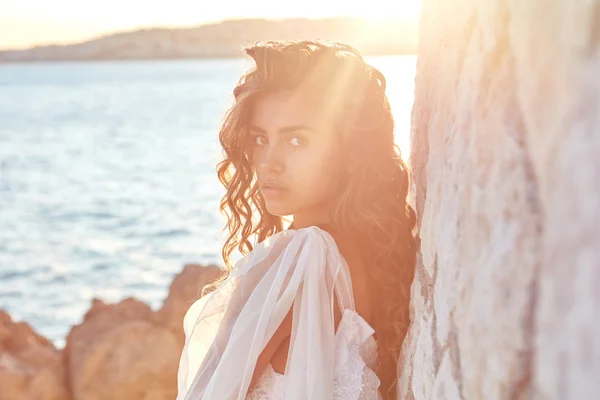 Retrato de bela sexy jovem modelo com cabelo escuro longo vestindo vestido branco longo posando perto de rocha no fundo do mar azul no verão — Fotografia de Stock