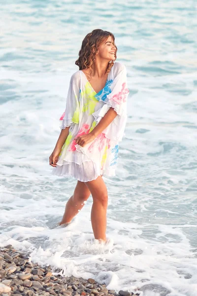 Modelo de mulher jovem sorridente muito sexy com cabelo escuro encaracolado longo vestindo vestido de praia andando ao longo da costa do mar deslumbrante no verão — Fotografia de Stock