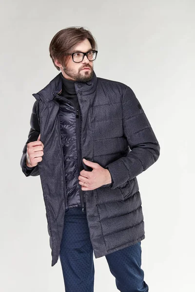 Attractive young sexy man model with beard and glasses posing in dark grey warm jacket and dark blue pants in light studio — Stock Photo, Image