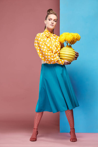 Pretty young serious woman model wearing yellow blouse with white polka-dot, blue skirt and pink tights, holding yellow vase with yellow flowers and posing in studio with blue and pink background