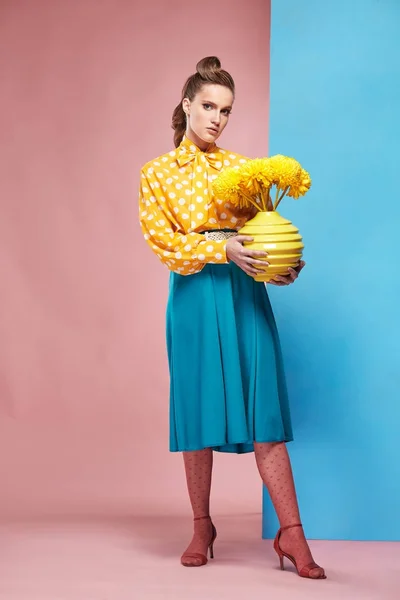 Beautiful young sexy woman model wearing yellow blouse with white polka-dot, blue skirt and pink tights in pin-up style, holding vase with yellow flowers and posing in studio with blue and pink background — Stock Photo, Image