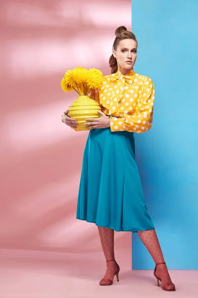 Hermosa modelo de mujer sexy joven con blusa amarilla con lunares blancos, falda azul y medias rosadas en estilo pin-up, sosteniendo jarrón con flores amarillas y posando en el estudio con fondo azul y rosa —  Fotos de Stock