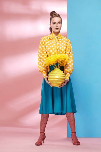 Beautiful young sexy woman model wearing yellow blouse with white polka-dot, blue skirt and pink tights in pin-up style, holding vase with yellow flowers and posing in studio with blue and pink background 