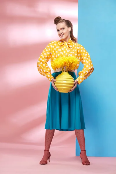 Increíble joven sexy mujer sonriente modelo con blusa amarilla con lunares blancos, falda azul y medias rosadas en estilo pin-up, sosteniendo jarrón con flores amarillas y posando en el estudio con fondo azul y rosa —  Fotos de Stock