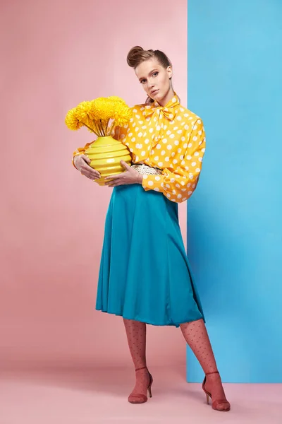 Όμορφη νεαρή γυναίκα σέξι μοντέλο φοράει κίτρινο μπλούζα με λευκό πουά, μπλε φούστα και ροζ κολάν σε στυλ pin-up, κρατώντας το βάζο με κίτρινα λουλούδια και θέτοντας σε στούντιο με μπλε και ροζ φόντο — Φωτογραφία Αρχείου