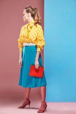 Pin-up stil, küçük kırmızı çanta tutarak ve Studio mavi ve pembe bir arka plan ile poz Beyaz benekli, mavi etek ve pembe tayt ile sarı bluz giyiyor büyük genç seksi ciddi kadın modeli 