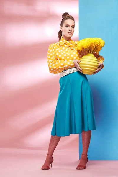 Increíble joven sexy mujer sonriente modelo con blusa amarilla con lunares blancos, falda azul y medias rosadas en estilo pin-up, sosteniendo jarrón con flores amarillas y posando en el estudio con fondo azul y rosa —  Fotos de Stock