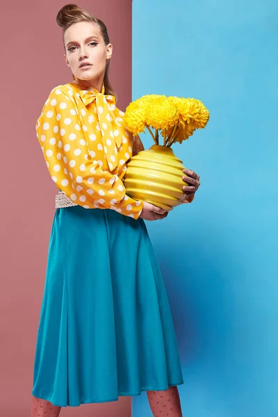 Gran modelo de mujer sexy joven con blusa amarilla con lunares blancos, falda azul y medias rosadas en estilo pin-up, sosteniendo jarrón con flores amarillas y posando en el estudio con fondo azul y rosa —  Fotos de Stock