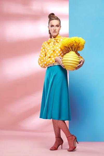 Modelo de mujer sexy bastante joven con blusa amarilla con lunares blancos, falda azul y medias rosadas en estilo pin-up, sosteniendo jarrón con flores amarillas y posando en el estudio con fondo azul y rosa —  Fotos de Stock