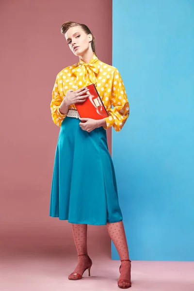 Bastante joven sexy modelo de mujer seria con blusa amarilla con lunares blancos, falda azul y medias rosadas en estilo pin-up, sosteniendo un pequeño bolso rojo y posando en el estudio con fondo azul y rosa —  Fotos de Stock